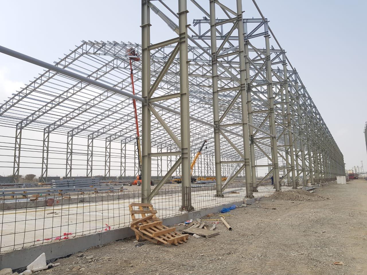 Workers assembling a steel frame section on a workshop floor under high ceiling industrial lighting